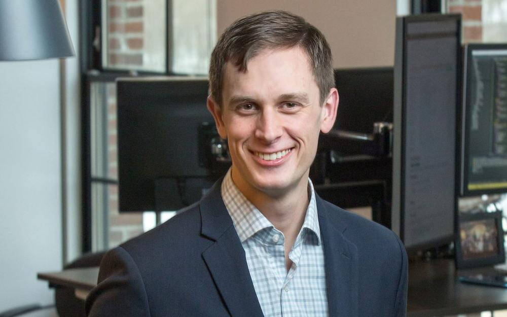A man stares right in to the camera. He is wearing a gray blazer and a checked shirt. Standing in front of computers.